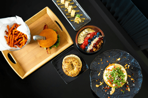 A variety of dishes including a burger, sweet potato fries, sushi, and a colorful fruit bowl on a black table.