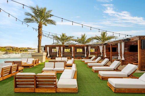Rooftop lounge area with wooden seating, white cushions, palm trees, and string lights under a clear blue sky.
