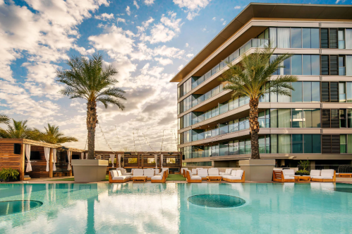 Modern hotel with a pool, palm trees, and lounge chairs under a blue sky with clouds.