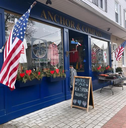 A storefront with "Anchor & Compass" sign, featuring winter sale signs, American flags, and spring arrivals displayed.