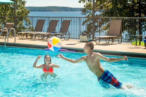 Two people play with a beach ball in a swimming pool, with a scenic lake and lounge chairs in the background.