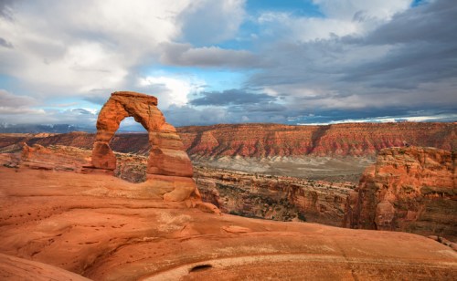 A stunning view of Delicate Arch in Arches National Park, surrounded by red rock formations and a cloudy sky.