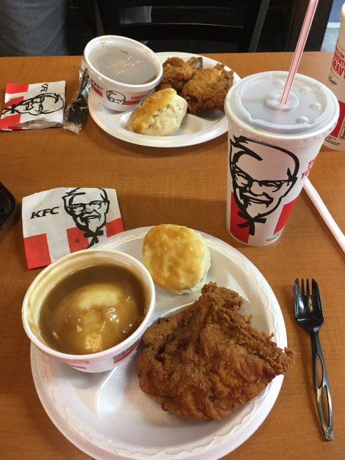 A KFC meal featuring fried chicken, mashed potatoes with gravy, a biscuit, and a drink on a wooden table.