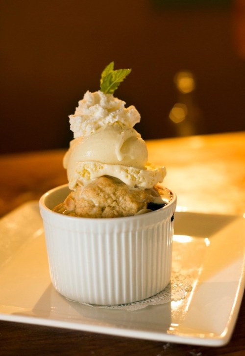 A warm dessert in a white ramekin topped with vanilla ice cream, whipped cream, and a mint leaf.