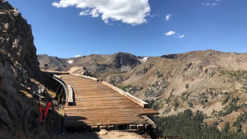 A wooden pathway curves along a mountain ridge, with a bike resting nearby and a clear blue sky overhead.