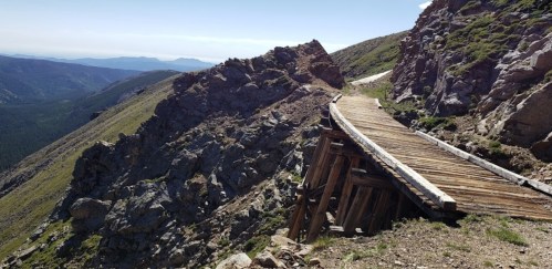 A wooden walkway winds along a rocky mountain slope, with distant peaks visible under a clear blue sky.