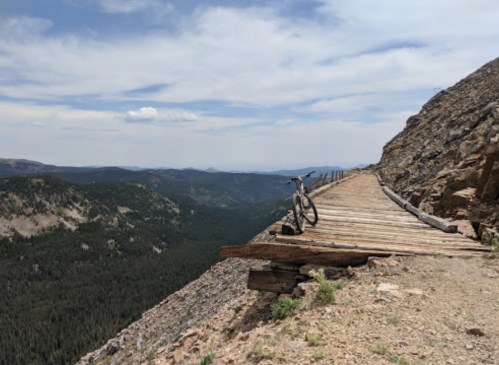 A mountain bike rests on a wooden path overlooking a vast, green valley and distant mountains under a blue sky.