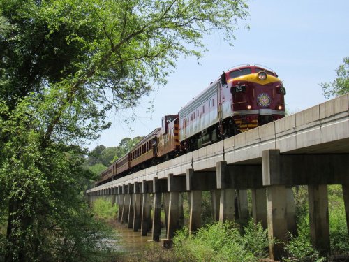 A vintage train crosses a bridge surrounded by lush greenery on a sunny day.