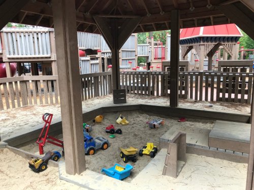 A sandbox area in a playground with various toy trucks and construction vehicles scattered on the sand.