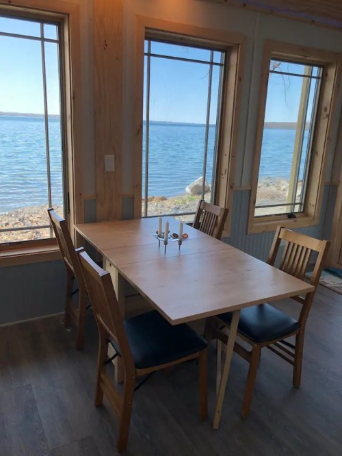 A wooden dining table with chairs by large windows overlooking a serene lake and clear blue sky.