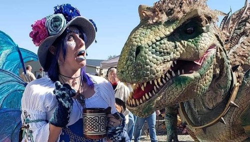 A woman in a colorful costume poses with a large dinosaur puppet, both looking surprised at each other.