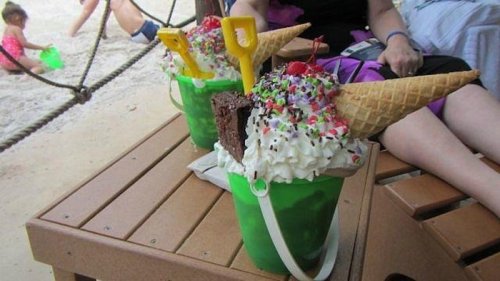 Two colorful ice cream sundaes in green buckets, topped with whipped cream, sprinkles, and cones, on a wooden table.