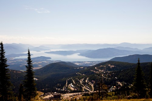 A panoramic view of mountains and a lake, with lush greenery and a clear blue sky in the background.