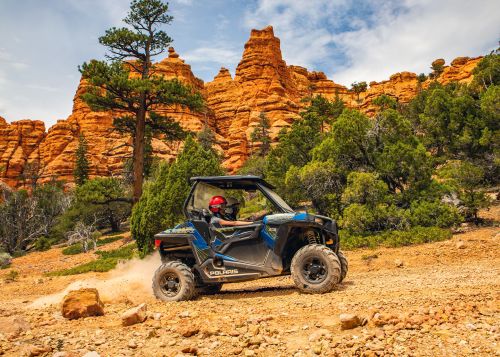 A blue off-road vehicle drives on a rocky trail with red rock formations and green trees in the background.