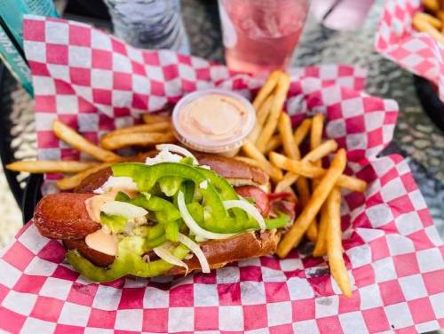 A hot dog topped with green peppers and onions, served with fries and a dipping sauce, on a red and white checkered paper.