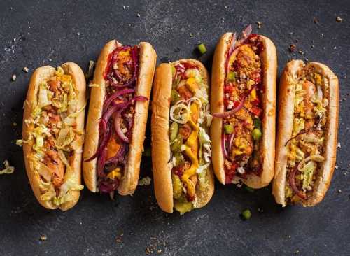 A variety of colorful hot dogs topped with different vegetables and sauces, arranged on a dark surface.