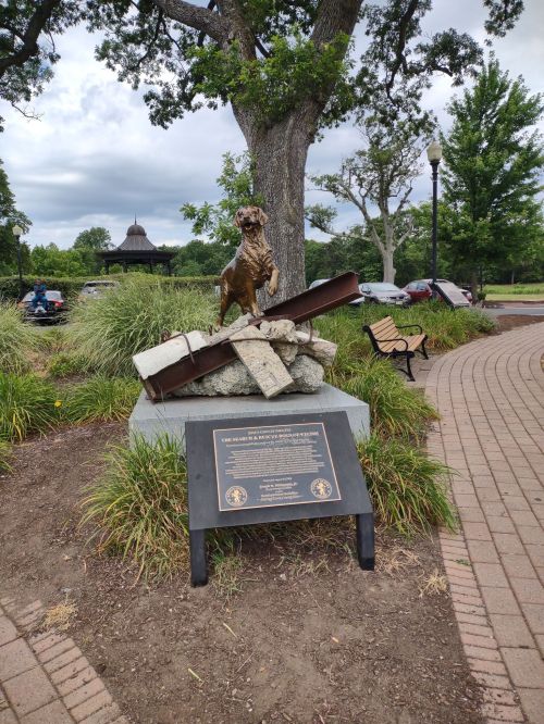 A bronze dog statue on a stone base, surrounded by greenery and a plaque, near a park path and benches.