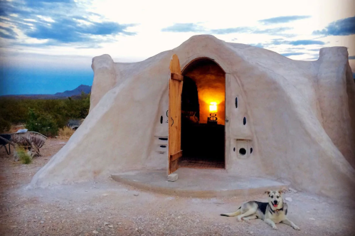 A unique adobe-style home at sunset, with a dog resting in front and mountains in the background.