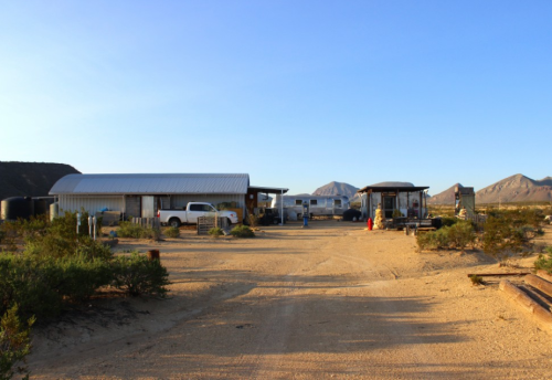A desert scene featuring several buildings and trailers along a dirt road, surrounded by sparse vegetation and mountains.