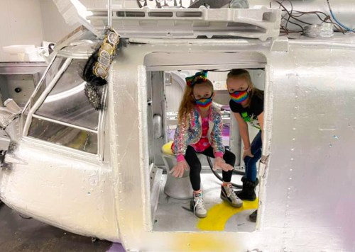 Two children in colorful masks sit inside a shiny, silver helicopter structure, smiling and posing for the camera.