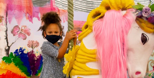 A child in a mask smiles while holding onto a colorful carousel horse with pink mane and vibrant decorations around.