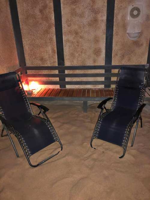 Two black lounge chairs on sandy ground, with a wooden bench and soft lighting in a cozy indoor setting.