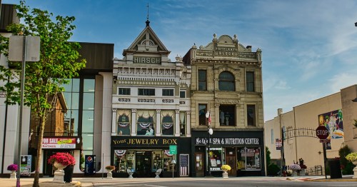 The historic downtown district of Wapakoneta, Ohio, showcases beautifully preserved brick buildings housing local businesses like "Your Jewelry Box" and "Be Well Spa," with decorative hanging flower baskets.