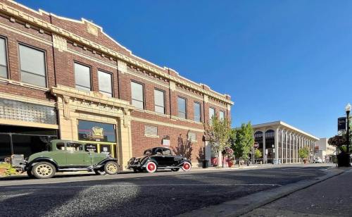 Two vintage cars parked on a street beside a historic brick building under a clear blue sky.