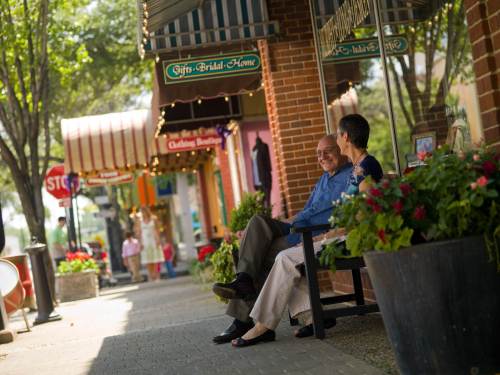 Two people sit on a bench outside shops, surrounded by greenery and flowers, enjoying a sunny day.