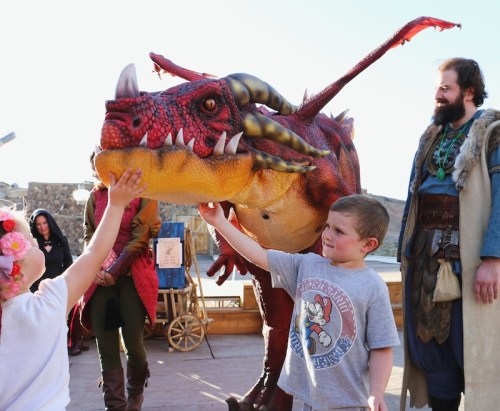 Children interact with a friendly dragon costume at a themed event, while an adult in medieval attire watches nearby.