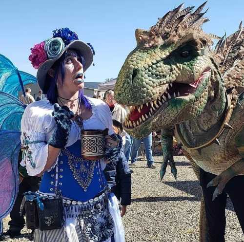 A person in a colorful costume stands beside a large, realistic dinosaur prop, both enjoying a lively outdoor event.