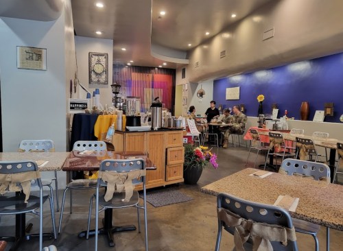 A cozy café interior with colorful walls, wooden tables, and patrons enjoying their meals. Flowers add a cheerful touch.
