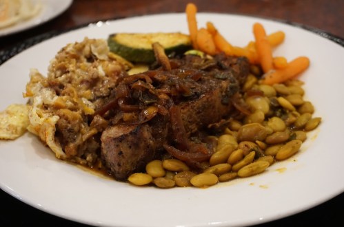 A plate of food featuring grilled meat, sautéed onions, lima beans, carrots, zucchini, and a side of baked dish.