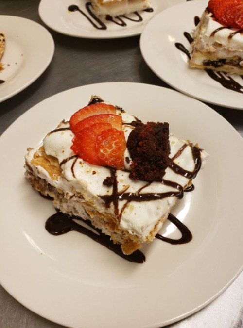 A slice of layered dessert topped with whipped cream, strawberries, and a brownie, drizzled with chocolate sauce.