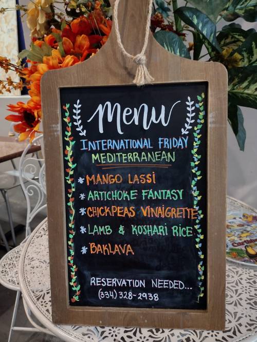 Chalkboard menu for International Friday featuring Mediterranean dishes: Mango Lassi, Artichoke Fantasy, Chickpeas Vinaigrette, Lamb & Koshari Rice, Baklava.