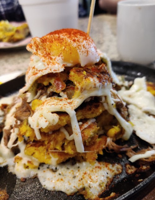 A stacked dish of cornbread, pulled meat, and a fried egg, drizzled with sauce and sprinkled with paprika.