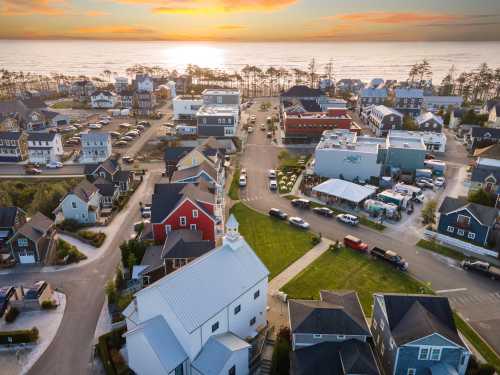 Aerial view of a coastal town at sunset, featuring houses, parked cars, and a glimpse of the ocean in the background.