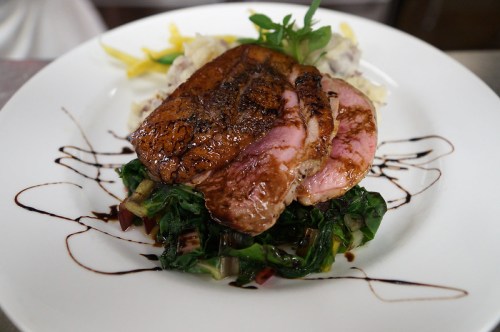 A plated dish featuring seared meat on a bed of greens, accompanied by mashed potatoes and drizzled with sauce.