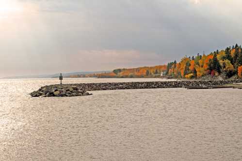 A tranquil lakeshore scene with a rocky pier and vibrant autumn foliage lining the distant shore under a cloudy sky.