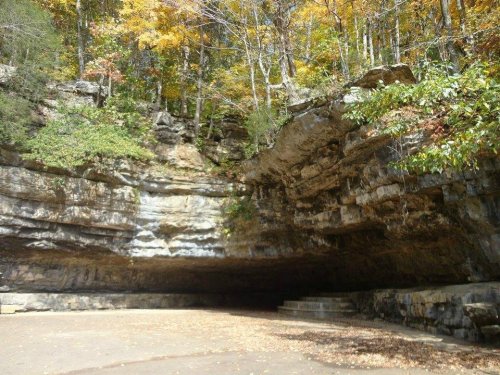 A rocky cave entrance surrounded by trees with autumn foliage, showcasing layered stone formations and a clear sky.