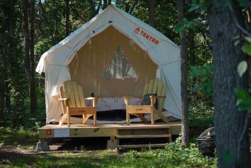 A cozy glamping tent with two wooden chairs on a wooden platform, surrounded by trees in a forested area.