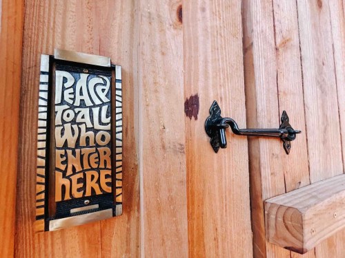 A wooden door with a brass plaque reading "Peace to all who enter here" and a decorative metal handle.
