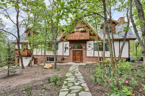 A charming multi-story house surrounded by trees and flowers, featuring a stone pathway and decorative wall art.