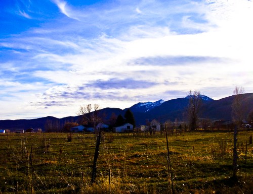 A scenic landscape featuring mountains, a blue sky with clouds, and a grassy field with a few houses in the distance.