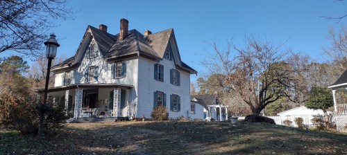 A large, white Victorian-style house with a steep roof, surrounded by trees and a clear blue sky.