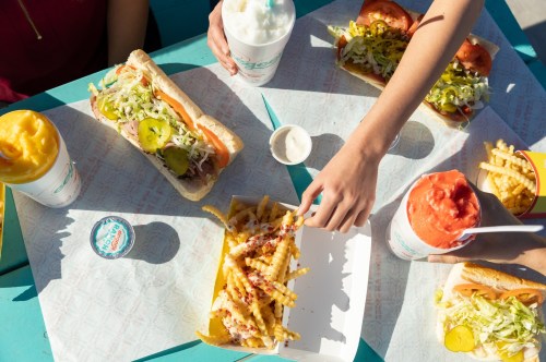 A colorful spread of hot dogs, fries, and drinks on a table, with hands reaching for the food.