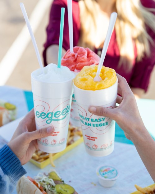 Three hands hold colorful Eegee cups, with a table of food in the background, celebrating with drinks.