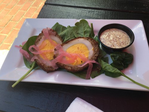 A plate with two sliced Scotch eggs on spinach, topped with pickled onions, and a side of mustard sauce.