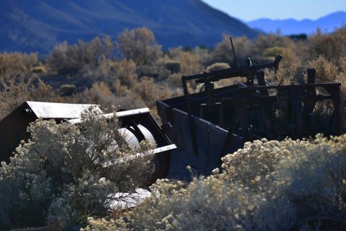 Rusty machinery partially covered by sagebrush, set against a backdrop of mountains and clear blue sky.
