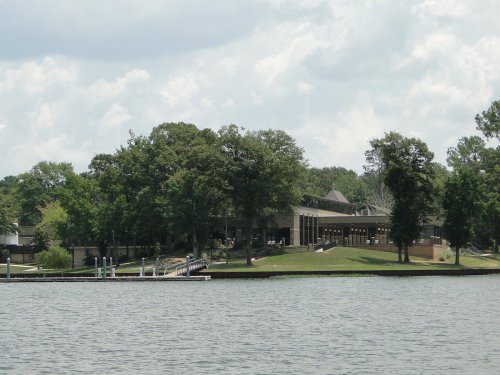 A lakeside view of a building surrounded by trees, with a dock extending into the water under a cloudy sky.
